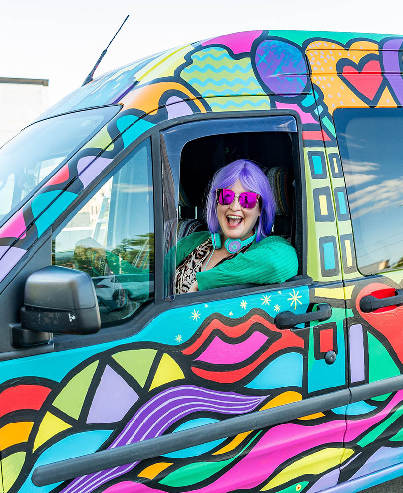 The host, Emily, driving the colorful van wearing a purple wig and pink sunglasses, smiling excitedly