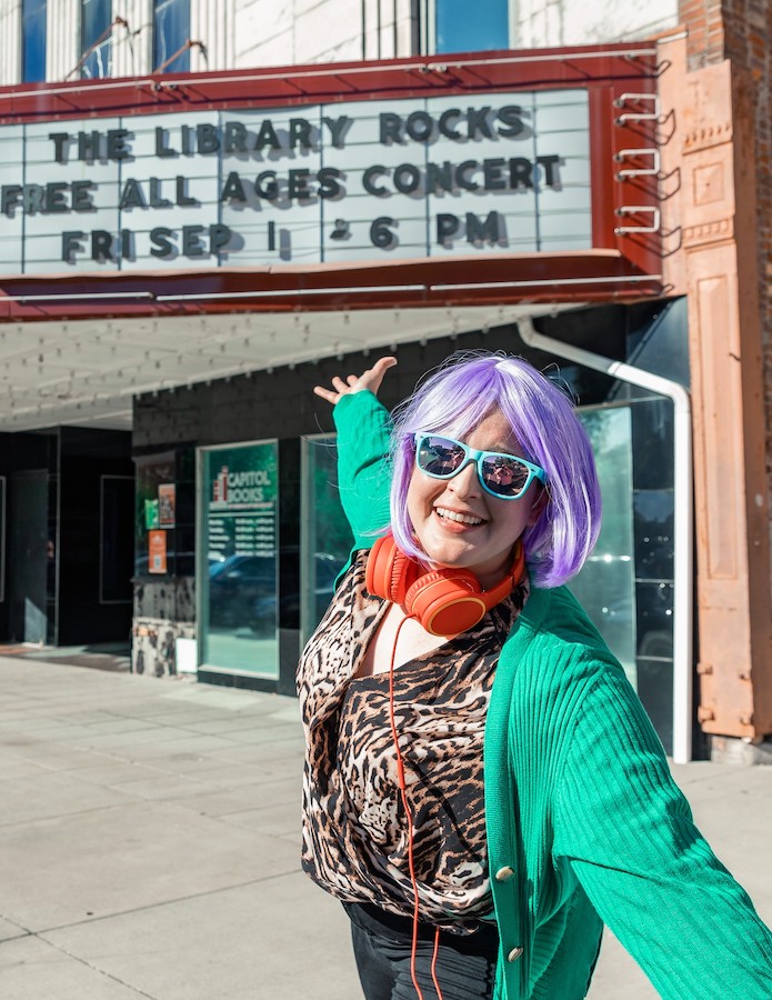 The host, Emily, in a purple wig and headphones in front of a marquee that says 'The Library Rocks - Free all ages concert Fri Sept 1'. Her arms are spread wide.