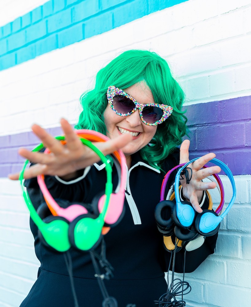 The host, Emily, smiling and wearing a green wig, holding several headphones in differing colors