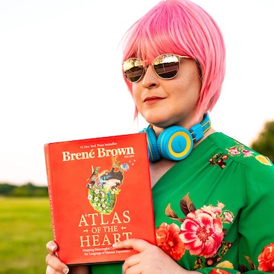 The host, Emily, wearing a pink wig, sunglasses and headphones, holding a book by Brene Brown